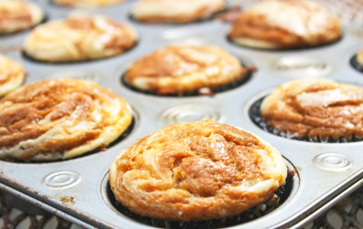 Close-up of a muffin tray filled with freshly baked, golden brown cinnamon swirl muffins. The muffins, inspired by pumpkin recipes, have a marbled pattern on top, and the tray is set on a cooling rack.