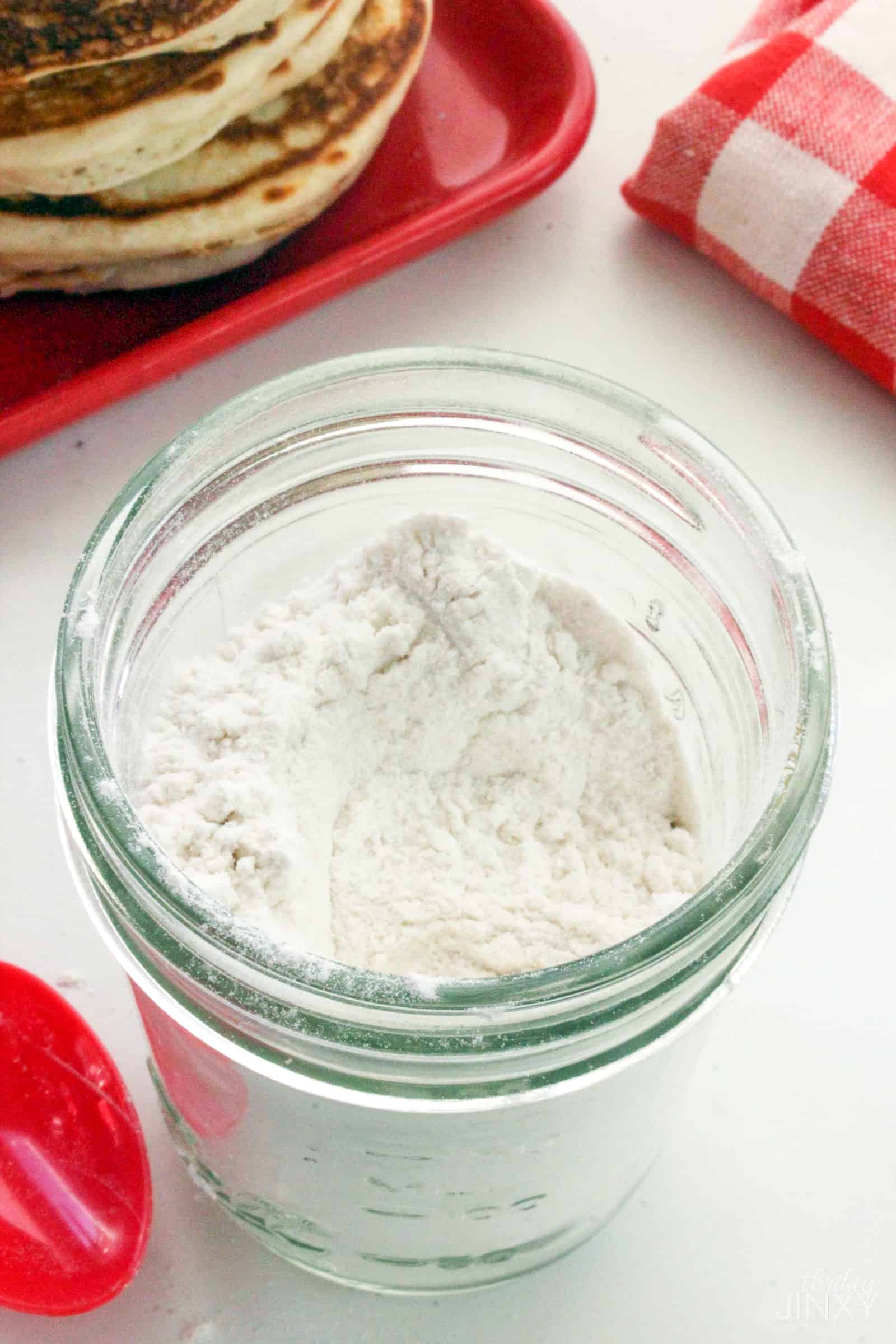 Glass jar with Homemade Pancake Mix next to a stack of pancakes and red checked towel.