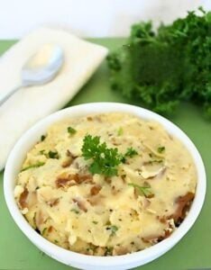 A bowl of creamy slow cooker garlic herb mashed potatoes garnished with parsley sits on a green surface. A spoon and a white napkin are placed nearby, with a bunch of fresh parsley in the background.