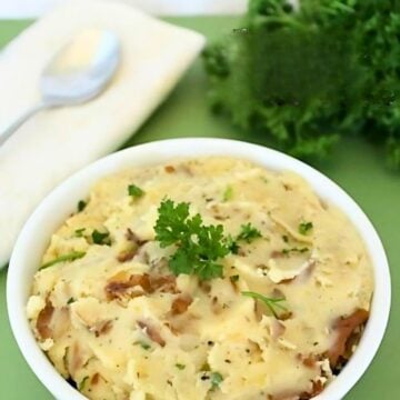 A bowl of creamy slow cooker garlic herb mashed potatoes garnished with parsley sits on a green surface. A spoon and a white napkin are placed nearby, with a bunch of fresh parsley in the background.