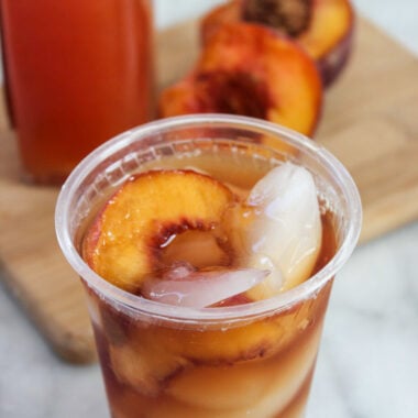 Copycat Sonic Peach Iced Tea in plastic cup with fresh peaches on cutting board in background