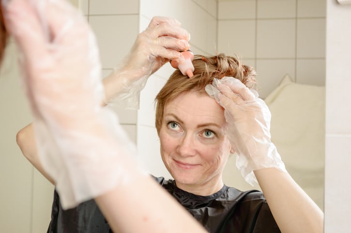 Caucasian woman with short hair dying her hair red in front of mirror in her own bathroom.