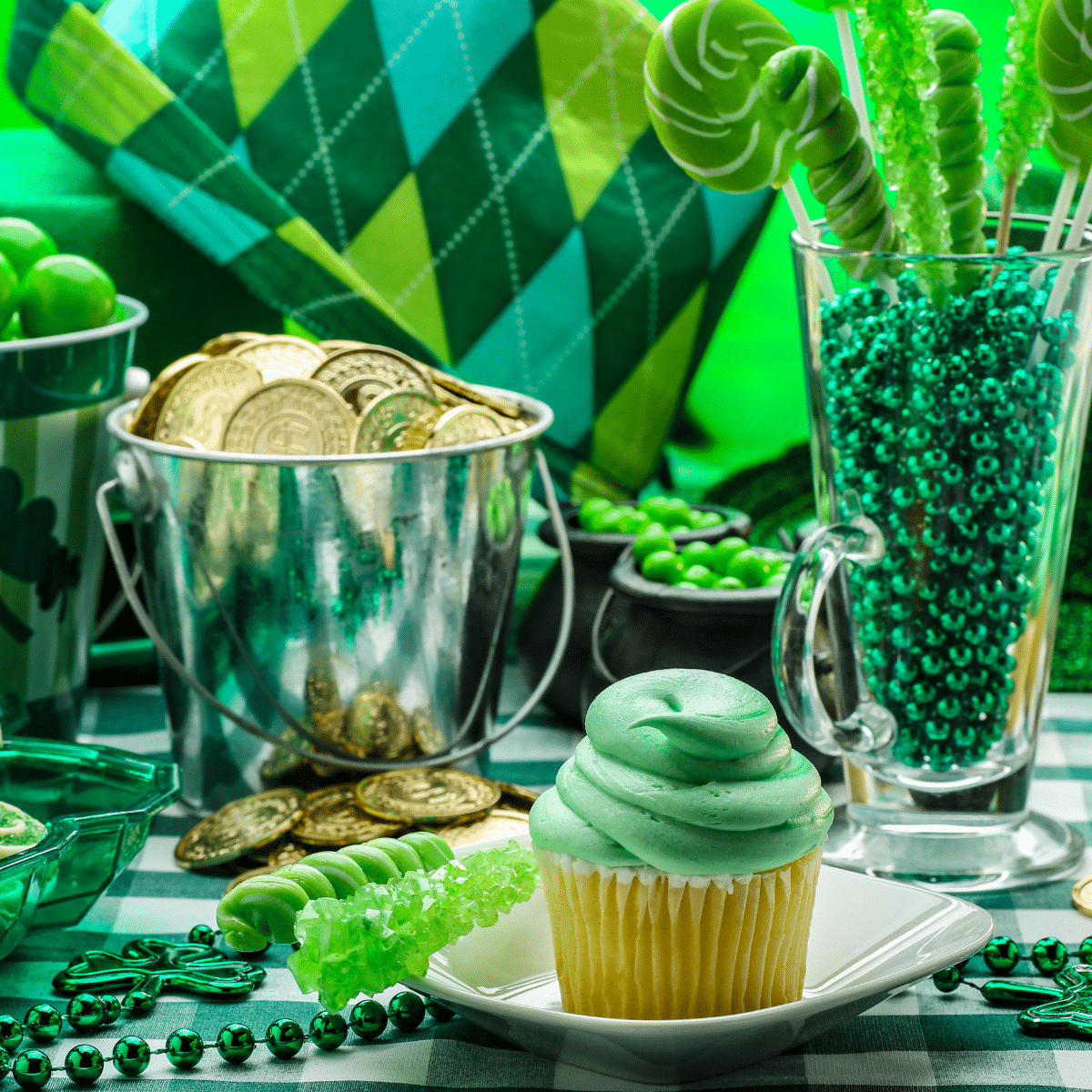 St Patrick's Day party table with green frosted cupcake, gold coins in a silver bucket, green beads and green candy.