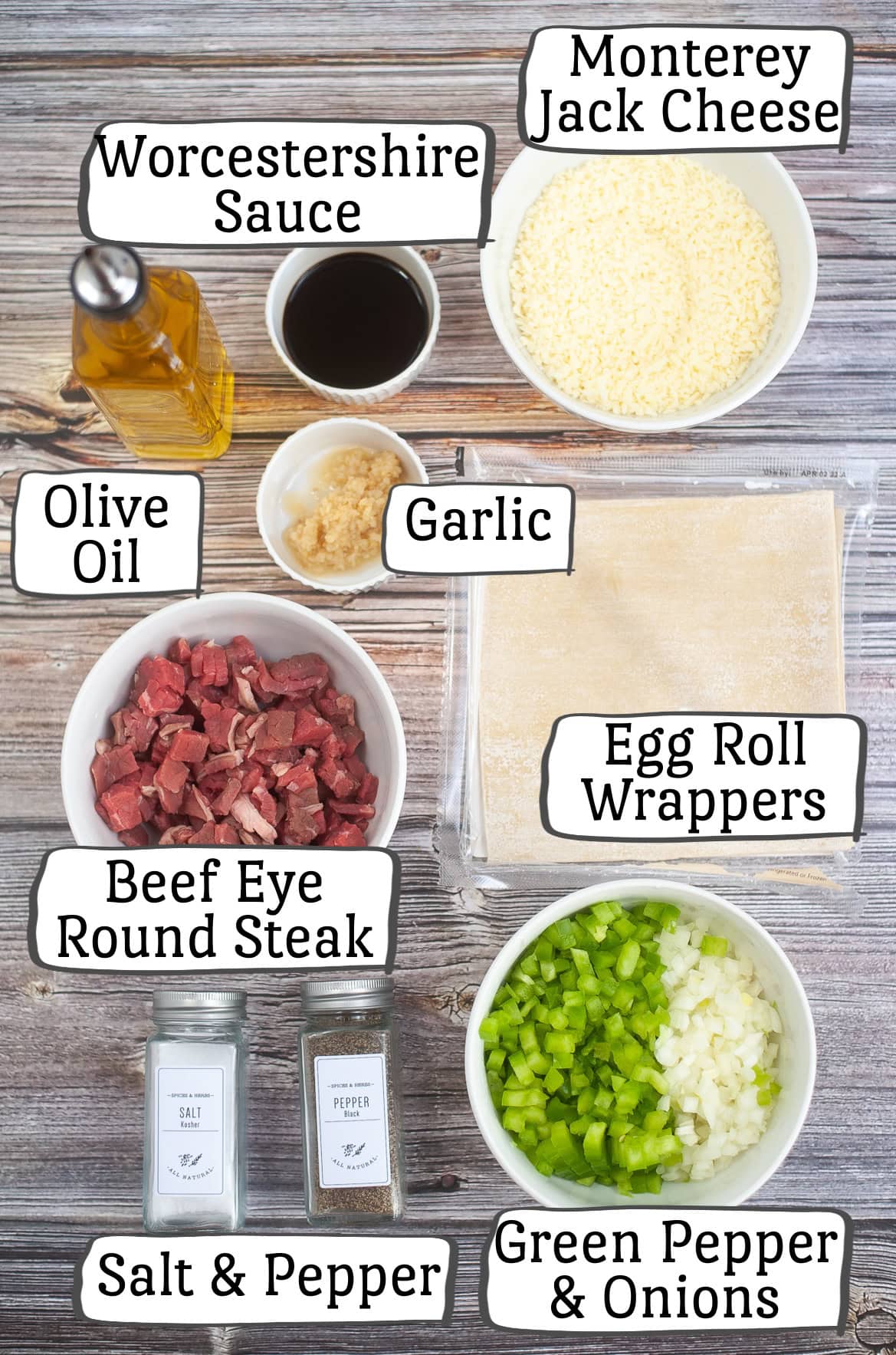 Ingredients for a recipe are laid out on a wooden surface, labeled in text: Worcestershire sauce, Monterey Jack cheese, olive oil, garlic, egg roll wrappers, beef eye round steak, salt & pepper, green pepper, and onions.