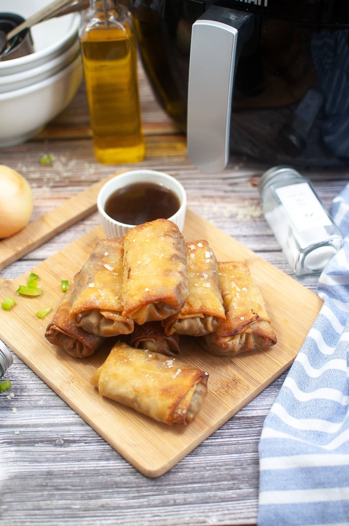 Crispy spring rolls stacked on a wooden board, served with a small bowl of dipping sauce. An air fryer, olive oil bottle, and onion are in the background, with a striped dish towel nearby.