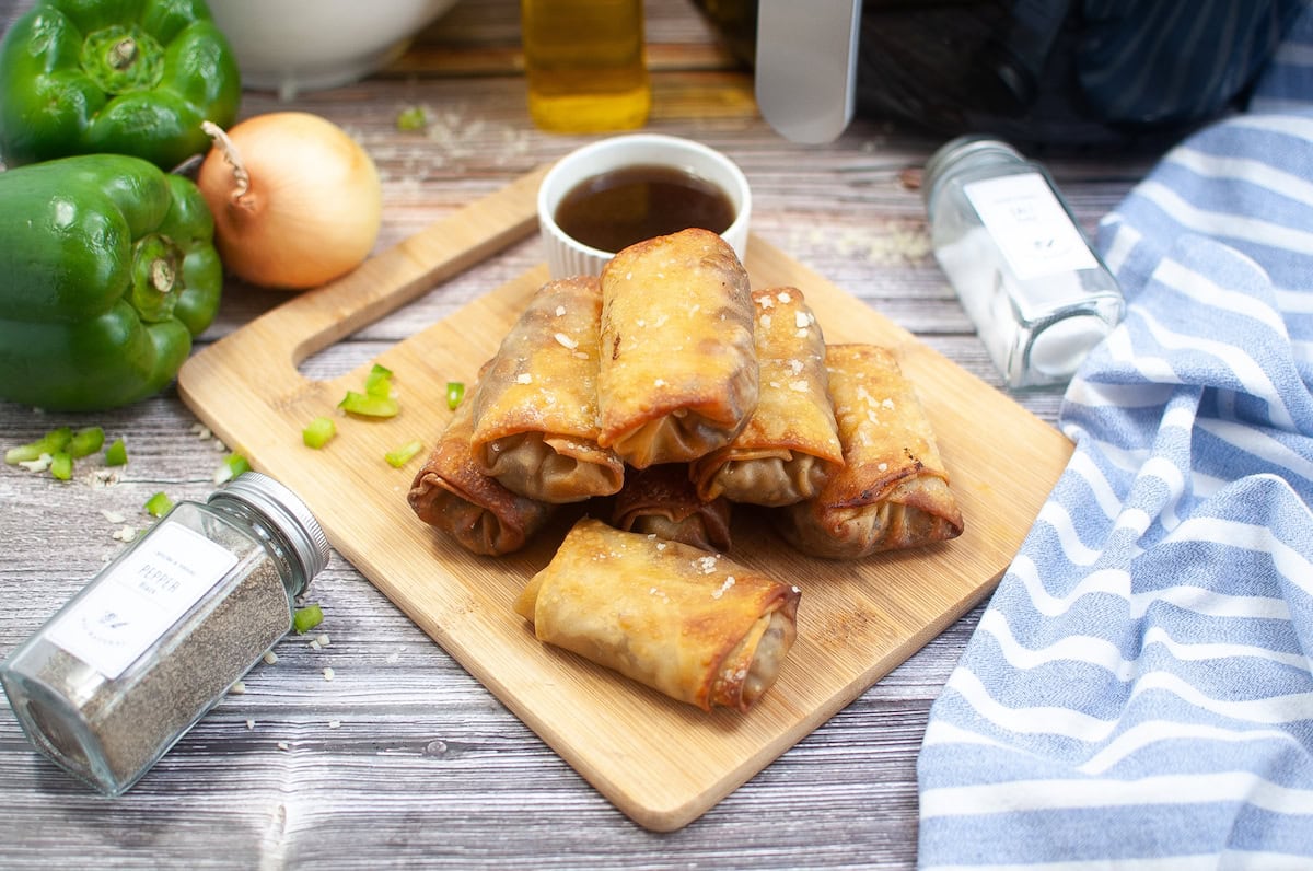 A wooden board topped with crispy golden philly cheesesteak eggrolls, accompanied by a small bowl of dipping sauce. Nearby are jars of spices, green bell peppers, an onion, and a striped cloth. The scene is set on a rustic wooden table.