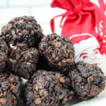 A plate of chocolate and cereal clusters is displayed with a red drawstring bag featuring a white Santa design. The background shows white tiles, and the plate rests on a black and white striped surface.