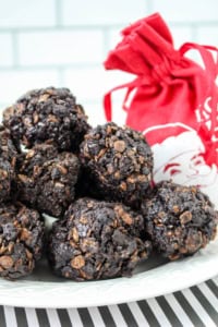 A plate of chocolate and cereal clusters is displayed with a red drawstring bag featuring a white Santa design. The background shows white tiles, and the plate rests on a black and white striped surface.