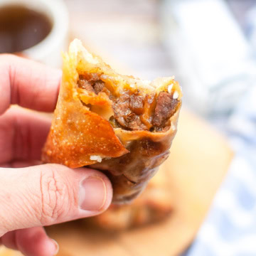 A hand holds a partially eaten, crispy fried egg roll filled with a savory beef mixture. A blurred background shows a cup of dipping sauce and a striped cloth on the table.