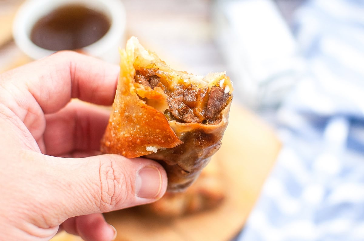 A hand holds a partially eaten, crispy fried egg roll filled with a savory beef mixture. A blurred background shows a cup of dipping sauce and a striped cloth on the table.