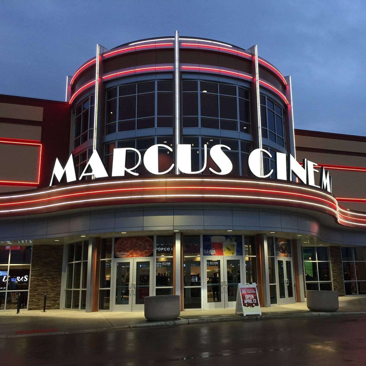 Front view of a modern movie theater with bright neon lights. The marquee displays "MARCUS CINEMA," and the building features glass windows and brick accents. The entrance includes poster displays and a wet pavement reflects the lights.