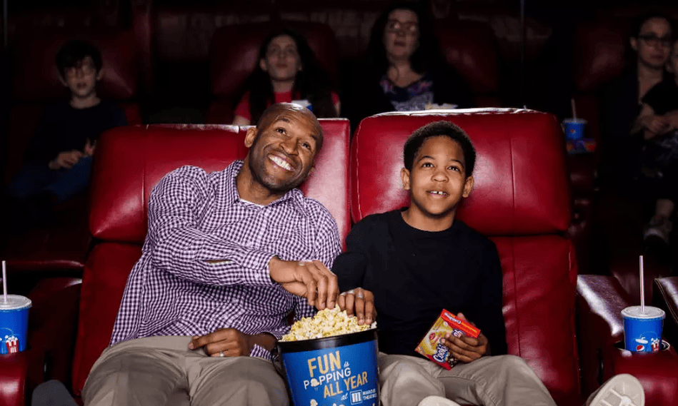 A man and a boy sit in a movie theater, enjoying popcorn together. They are seated on red reclining chairs, surrounded by other moviegoers. The theater is dimly lit, and both appear engaged and happy while watching the screen.