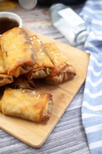 A stack of crispy, golden-brown egg rolls on a wooden cutting board next to a blue and white striped cloth. A small dish with dipping sauce and an upside-down spice bottle are in the background.