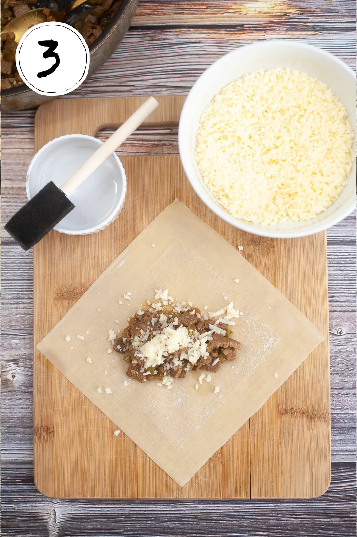 A wooden board with a sheet of dough containing a filling of chopped cooked meat and grated cheese. A bowl filled with more cheese, a small brush, and a dish are nearby. A white circle with the number "3" is in the top left corner.