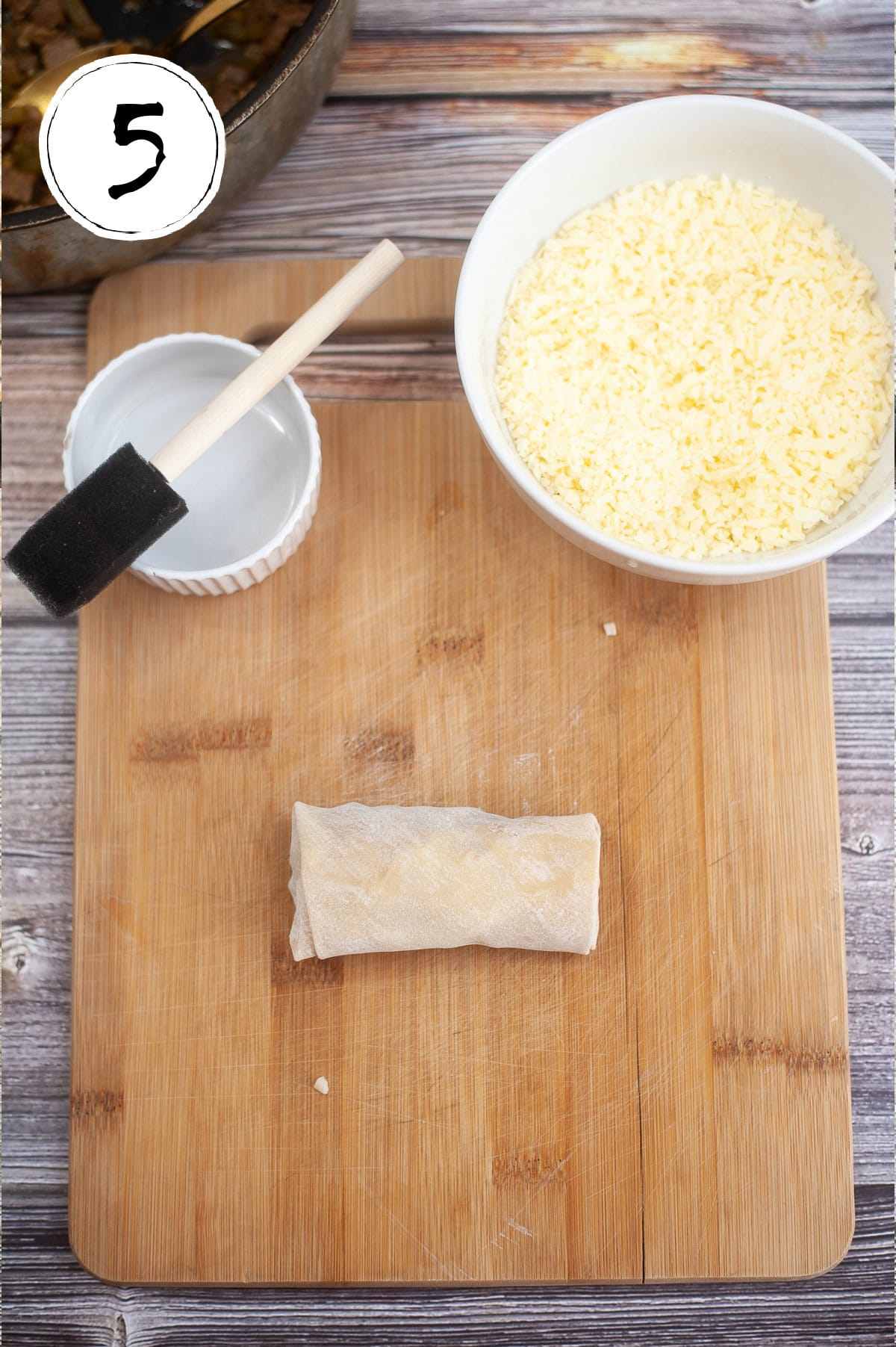 A wooden cutting board with a small wrapped pastry in the center. To the side, a bowl filled with shredded cheese and a small dish with a black foam brush are visible. The number 5 is in the top corner.