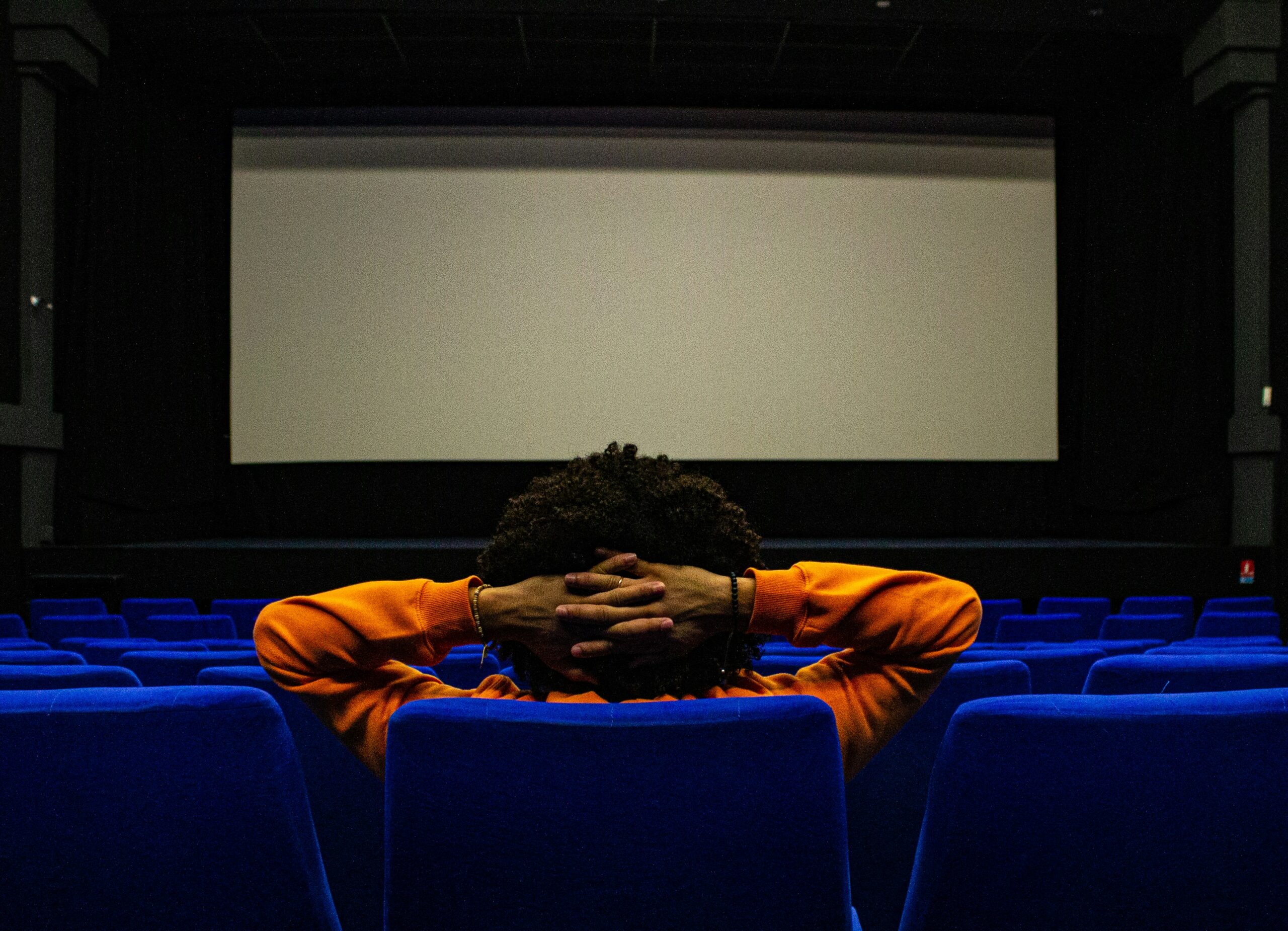A person in an orange shirt relaxes with their hands behind their head, sitting alone in a theater with blue seats, facing an empty white screen.
