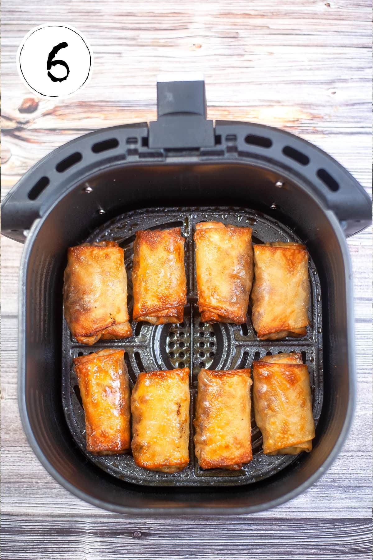Eight golden brown spring rolls are arranged neatly in an air fryer basket. The background features a wooden surface, and the number "6" is written in the top left corner.