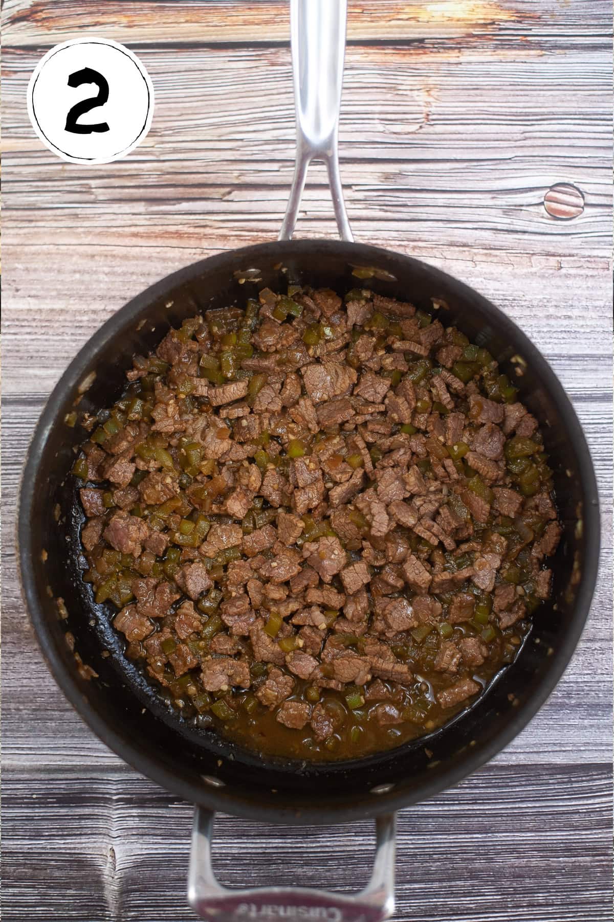 A pot filled with chopped, cooked beef and green bell peppers on a wooden surface. The number "2" is shown inside a white circle in the top left corner.