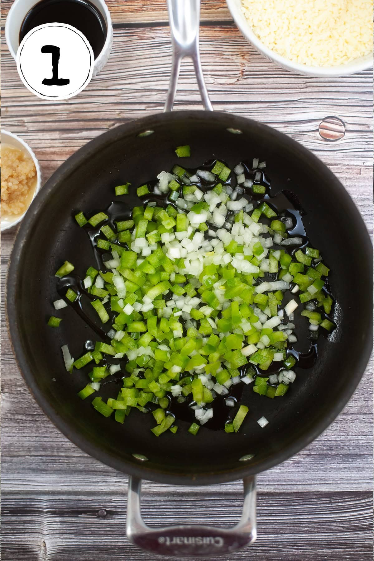A pot on a wooden surface contains chopped green bell peppers and onions. Nearby, there's a small bowl of minced garlic, another bowl with dark liquid, and a bowl of grated cheese. A white circle with the number 1 is in the top left corner.