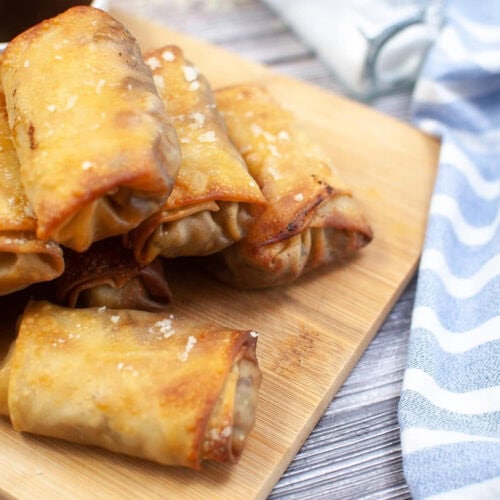 A stack of crispy, golden-brown egg rolls on a wooden cutting board next to a blue and white striped cloth. A small dish with dipping sauce and an upside-down spice bottle are in the background.