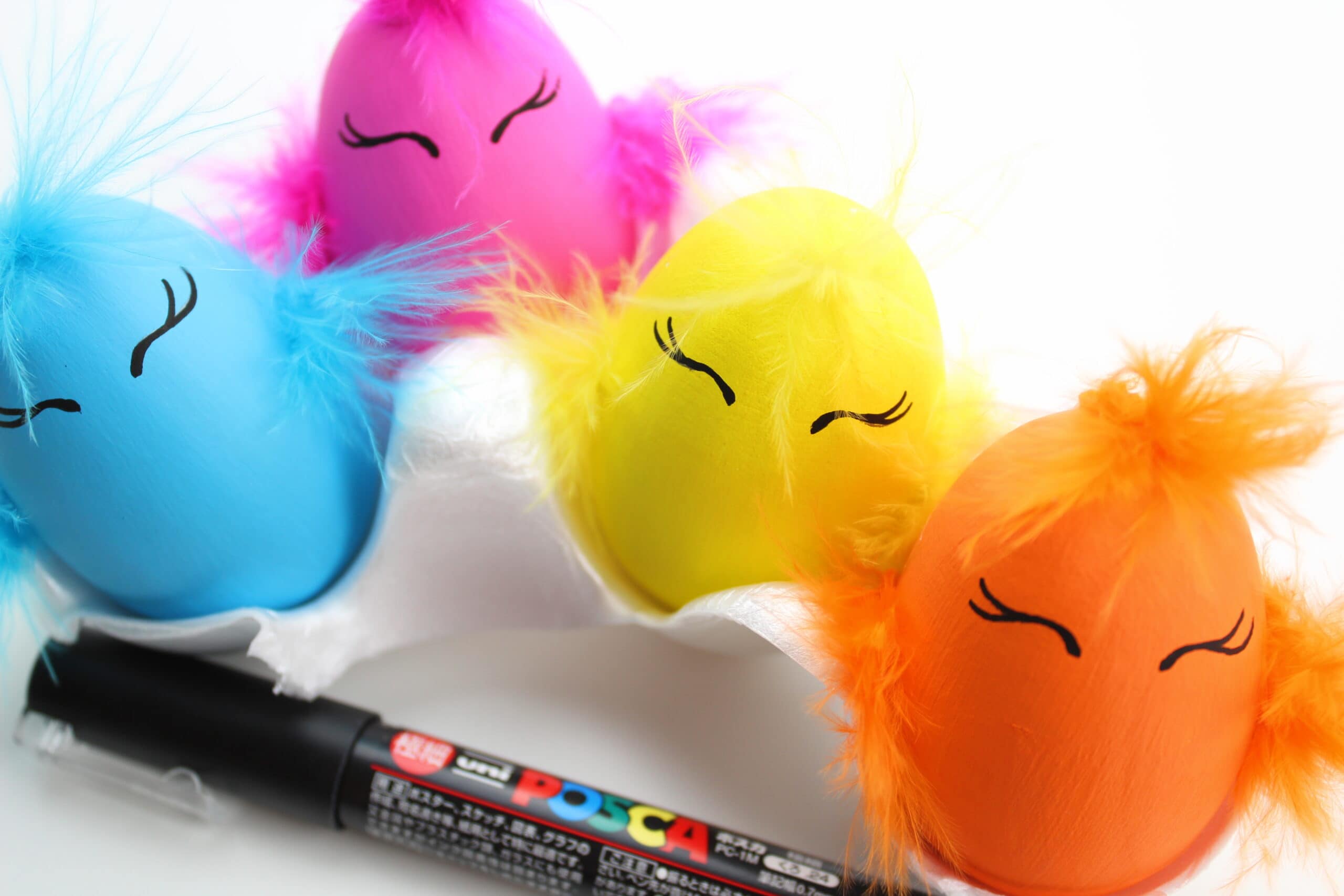 Brightly painted easter eggs with feathers glued on and eyes made with black marker. The black marker is in front of the eggs.