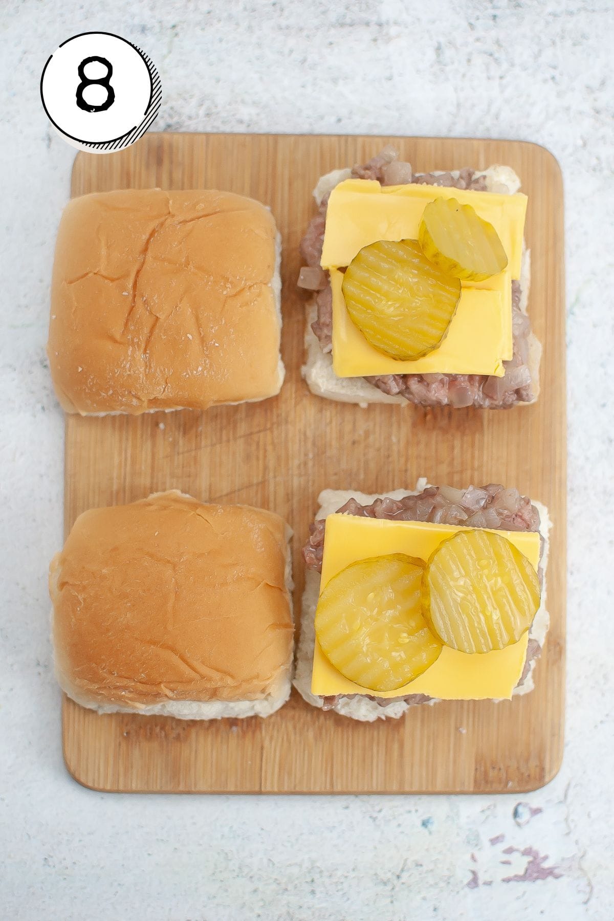 Assembling the sliders with a bottom bun, then a burger patty, slices of cheese cut to fit the patties, and 2 pickles on top; topping with other half of bun.