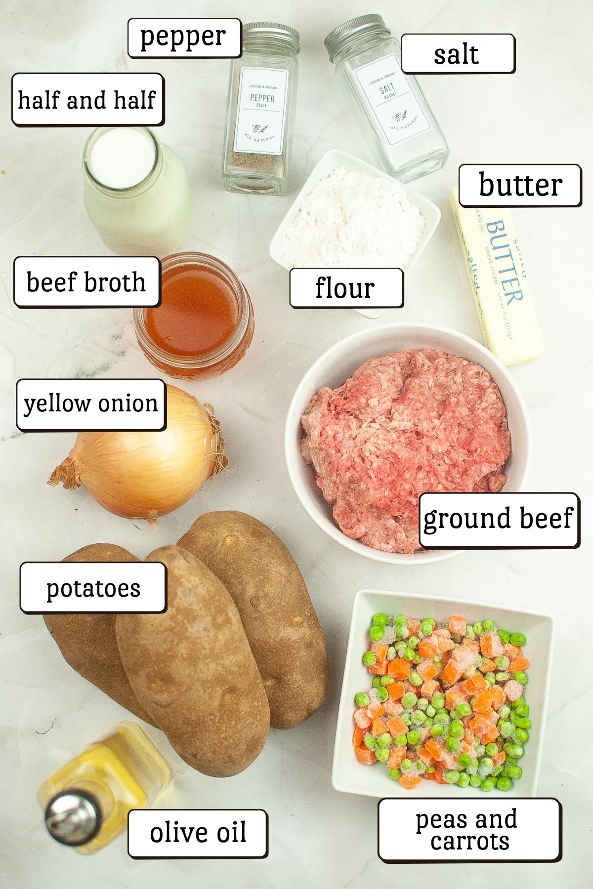 Overhead view of labeled ingredients for air fryer shepherd&rsquo;s pie baked potatoes, including ground beef, potatoes, peas and carrots, yellow onion, beef broth, flour, butter, half and half, olive oil, salt, and pepper.