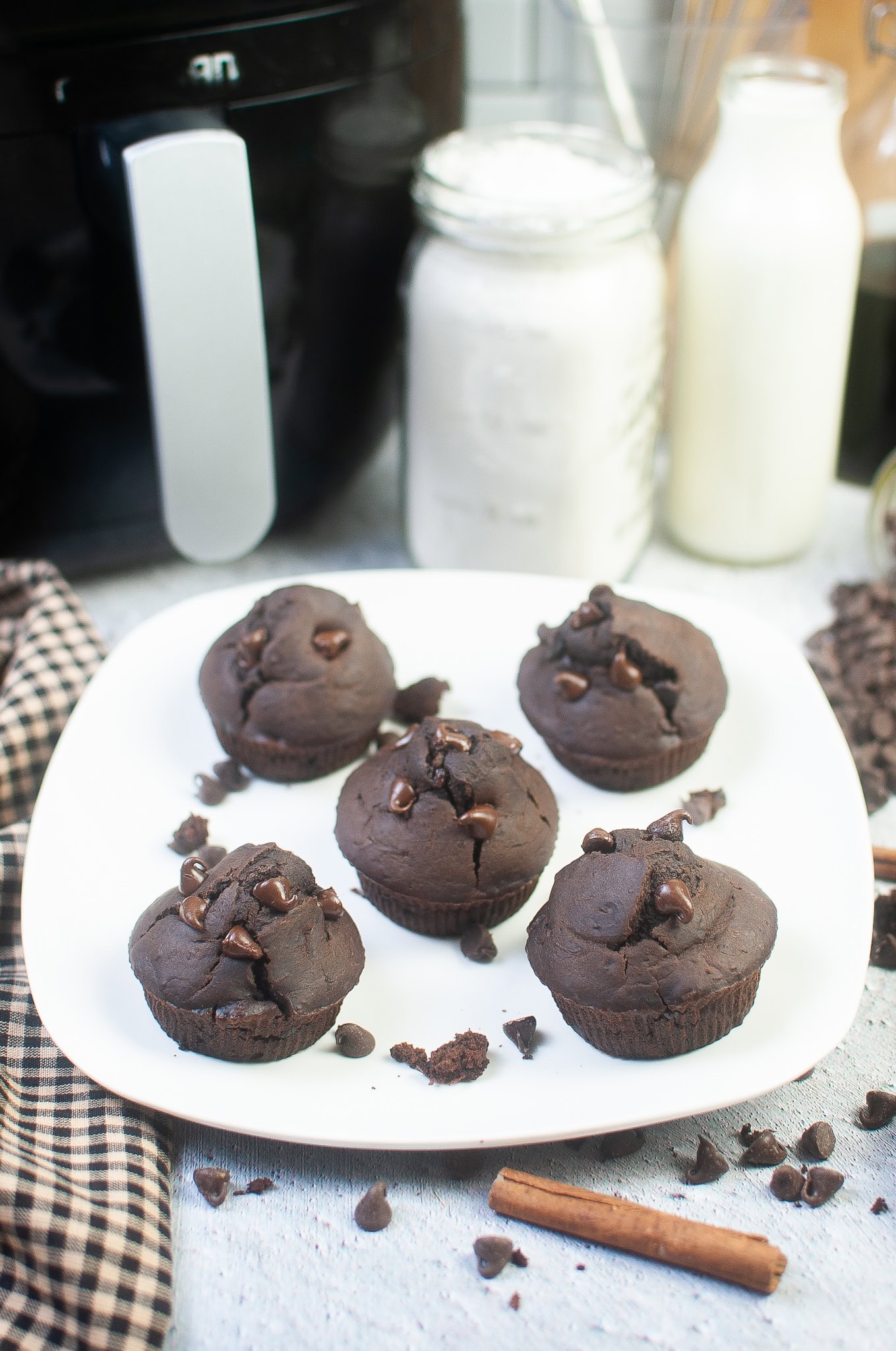 Plate of double chocolate zucchini muffins topped with chocolate chips, styled with an air fryer, milk bottles, and baking ingredients in the background.