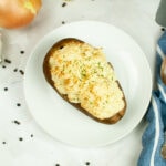 Overhead view of a shepherd’s pie baked potato topped with golden mashed potatoes and parsley, served on a white plate with a spoon and blue striped napkin.