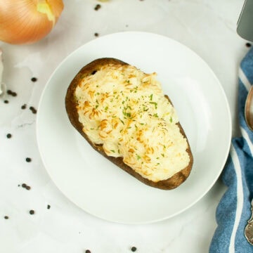 Overhead view of a shepherd's pie baked potato topped with golden mashed potatoes and parsley, served on a white plate with a spoon and blue striped napkin.