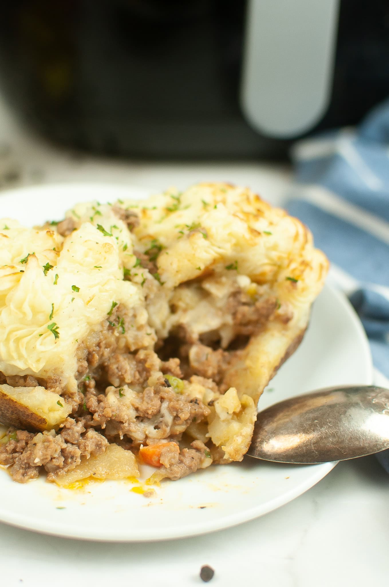 Close-up of a baked potato filled with shepherd&rsquo;s pie, showing seasoned ground beef, vegetables, and creamy mashed potato topping, served on a white plate with a spoon.