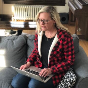 Woman with blond hair and buffalo plaid jacket using laptop on sofa.